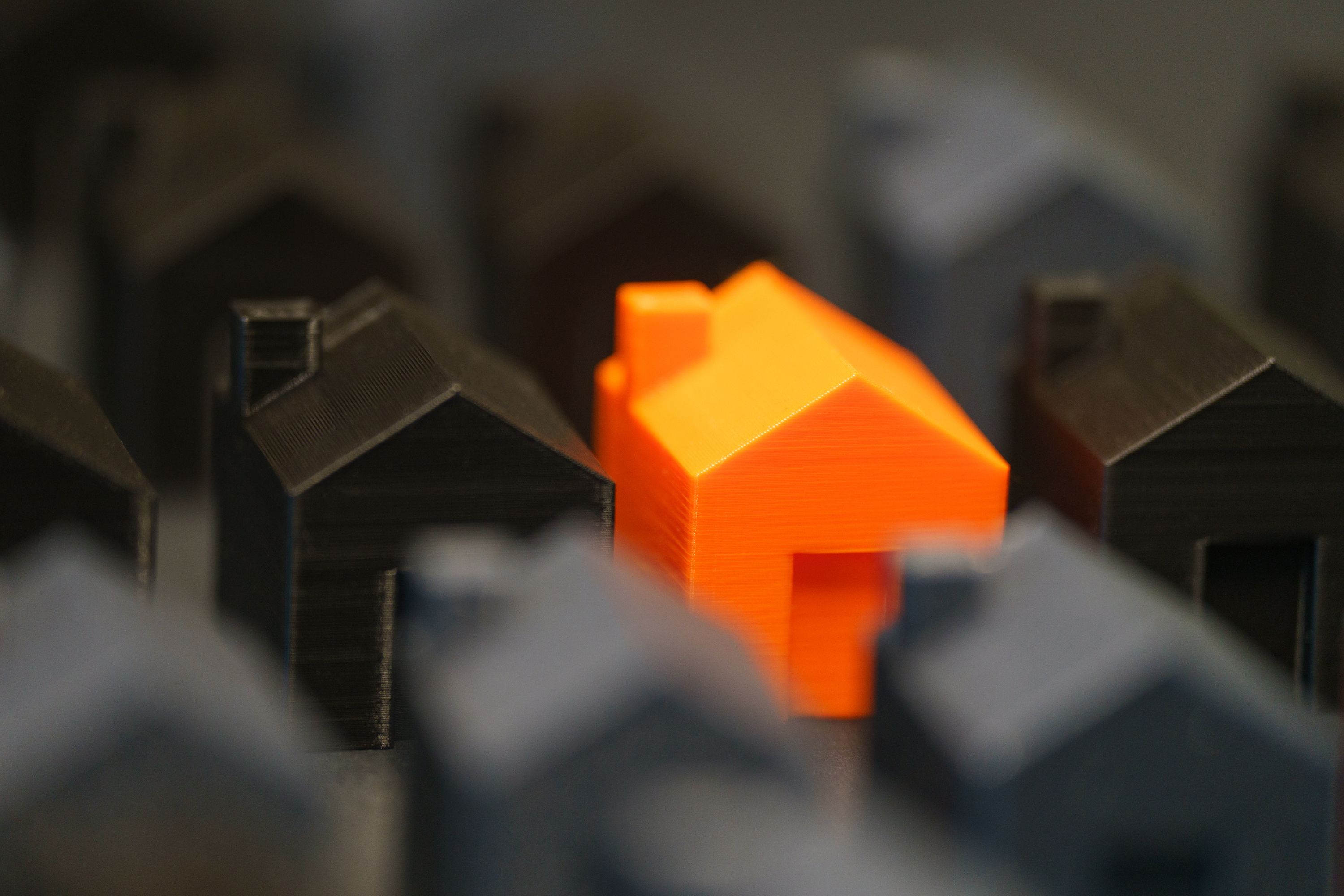 A vibrant orange model house standing among black miniature houses, symbolizing housing affordability as a local voting issue.